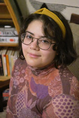 Close up portrait of a light-skinned woman, with short black hair, wearing a yellow headband and a multi-colored pink long sleeve blouse