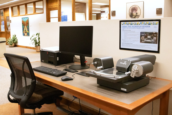 a black PC desktop, monitor, keyboard, mouse, and microfilm reader sitting atop a gray and brown table, with a chair tucked under the table