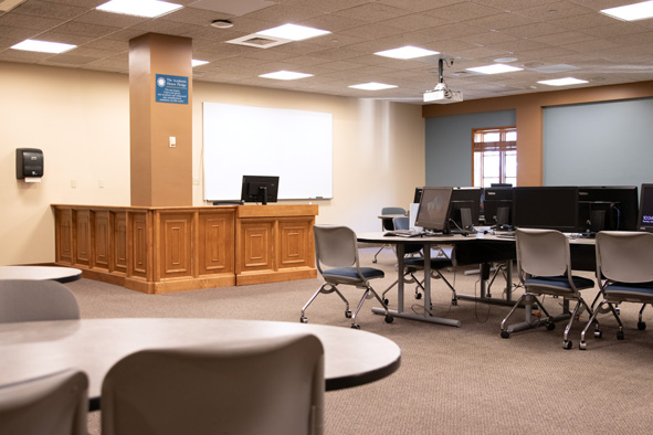 PC desktops and gray chairs with blue cushions banked at long tables, facing an -shaped podium in front of a dry erase board