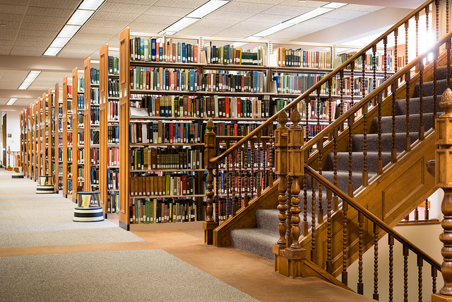 Sunlight shining through a window behind bookshelves filled with books behind a wooden staircase